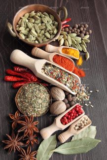 عکس با کیفیت هل سبز داخل ظرف سفالی و ادویه جات داخل پیمانه های چوبی و فلفل قرمز و چند برگ و لیمو بر روی میز چوبی