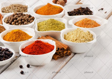 عکس با کیفیت پودر زنجبیل و پودر فلفل قرمز و زرد چوبه و میخک و فلفل شیرین و چند ادویه دیگر داخل پیاله های چینی سفید بر روی میز چوبی