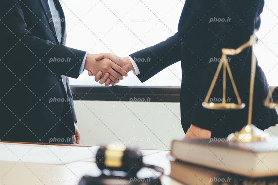 عکس با کیفیت وکیل و موکل در حال دست دادن و ترازو عدالت و چکش عدالت بر روی میز