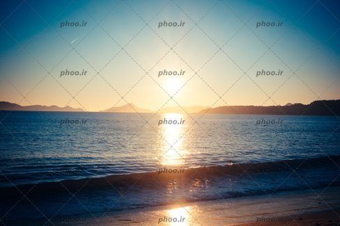 عکس با کیفیت طلوع افتاب و انعکاس نور خورشید بر روی آب و صخره ها و کوه ها در دور دست ها
