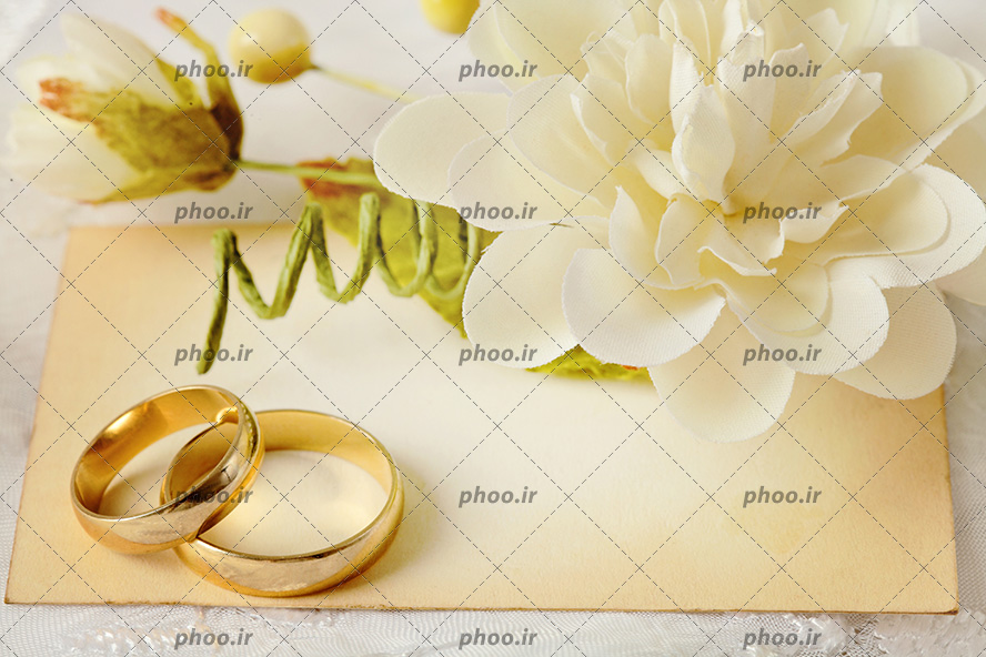 عکس با کیفیت دو حلقه طلایی بر روی کارت طلایی رنگ و گل زیبا به رنگ سفید در کنار حقله ها