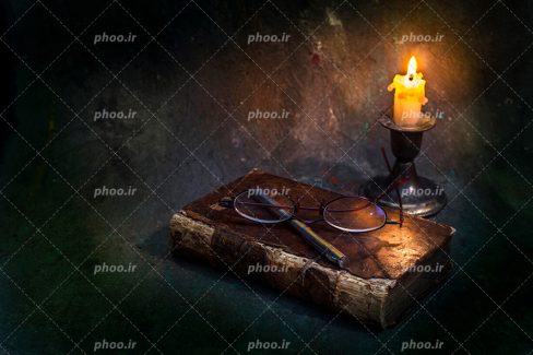 عکس با کیفیت شمع در شمعدانی در کنار کتاب و عینک مطالعه