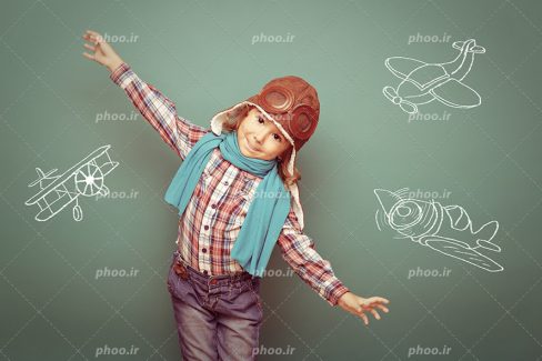عکس با کیفیت کودک با لباس خلبانی و گرفتن دست های خود به شکل بال های هواپیما و پس زمینه تخته سیاه با نقاشی هواپیماها