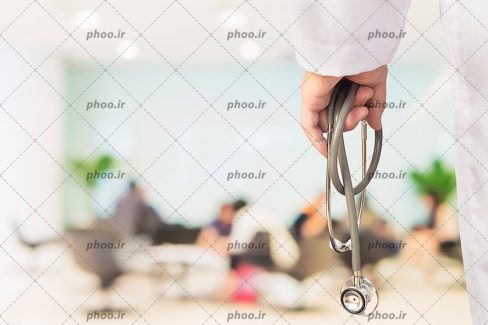 عکس با کیفیت استتوسکوپ در دست پزشک از نمای نزدیک و پس زمینه محو از بیمار های نشسته بر روی صندلی های انتظار