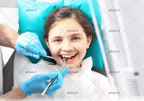 عکس با کیفیت کودک زیبا با چشم های آبی نشسته بر روی صندلی در دندانپزشکی و دندانپزشک در حال برسی و درست کردن دندان های دختر بچه