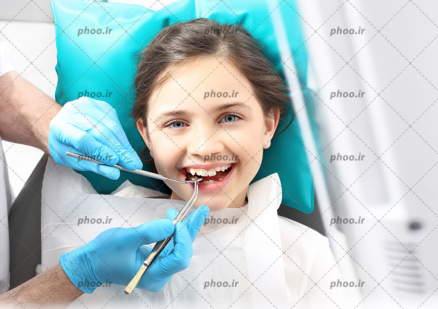 عکس با کیفیت کودک زیبا با چشم های آبی نشسته بر روی صندلی در دندانپزشکی و دندانپزشک در حال برسی و درست کردن دندان های دختر بچه