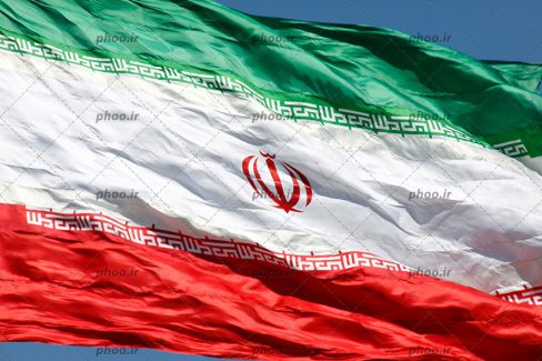 عکس با کیفیت آسمان به رنگ آبی و پرچم ایران در حال تکان خوردن به دلیل وزش باد