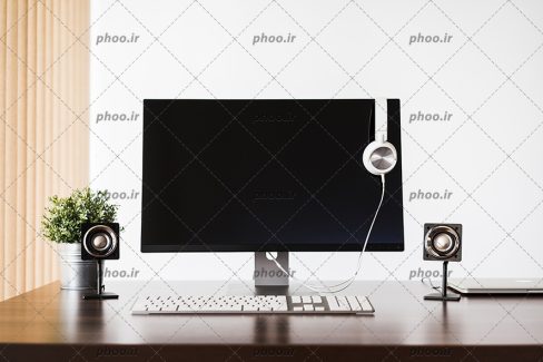 عکس با کیفیت مانیتور و کیبورد بر روی میز کامپیوتر و دو باند کوچک در کنارش و هدفون بر روی مانیتور کامپیوتر