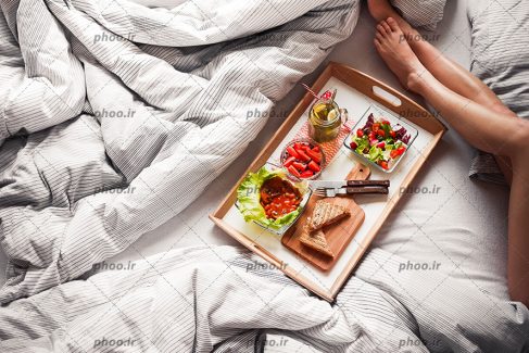 عکس با کیفیت سینی صبحانه در تخت خواب و پاهای زن در گوشه تصویر