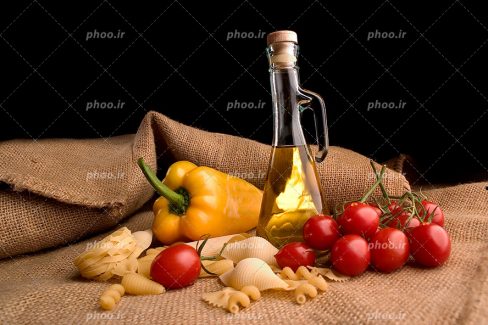 عکس با کیفیت انواع پاستا ها در کنار شیشه روغن و تعدادی گوجه و فلفل دلمه ای زرد بر روی پارچه ای کنفی