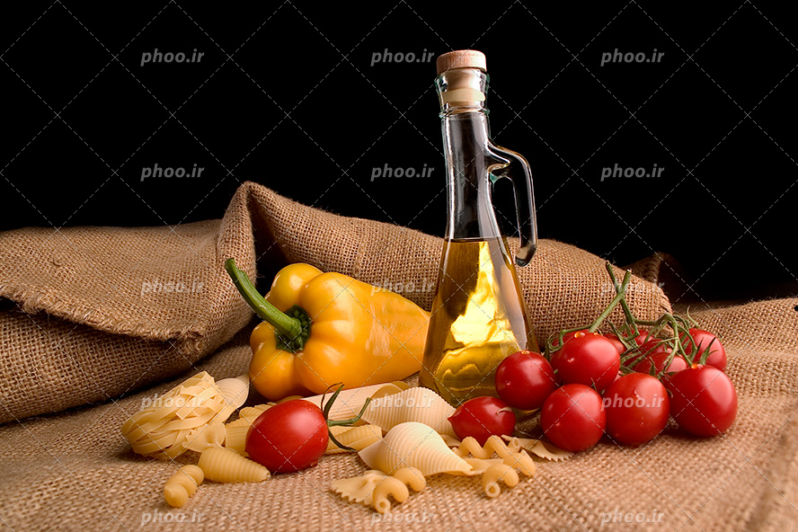 عکس با کیفیت انواع پاستا ها در کنار شیشه روغن و تعدادی گوجه و فلفل دلمه ای زرد بر روی پارچه ای کنفی