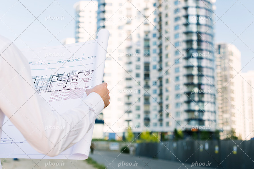 عکس با کیفیت مهندس عمران با لباسی سفید در حال نگاه کردن به نقشه در مقابل ساختمان