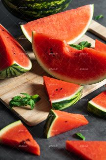 عکس با کیفیت هنوانه ی نصف شده در کنار چند اسلایس قرار گرفته بر روی تخته گوشت