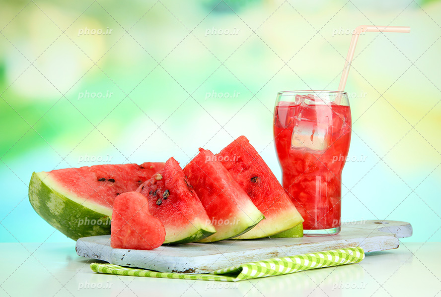 عکس با کیفیت پارچه چهارخونه سبز در زیر سنگ و آب هندوانه بر روی آن و قاچ های قلبی شکل و مثلثی هندوانه در کنار آن
