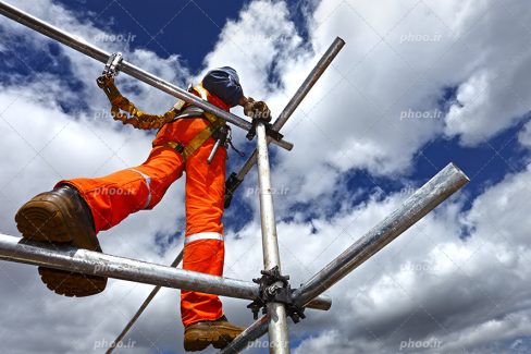 عکس با کیفیت کارگر بر روی داربست با کفش های ایمنی و لباس ایمنی و کلاه ایمنی با پس زمینه آسمان ابری
