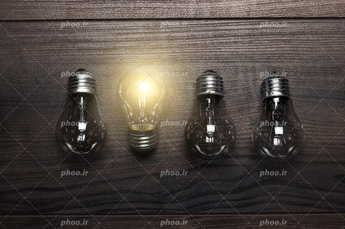 عکس با کیفیت زیبا یک لامپ روشن در بین لامپ های خاموش قرار گرفته بر روی میز چوبی