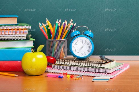 عکس با کیفیت سیب در کنار دفاتر و کتاب های رنگارنگ و ساعت زنگدار بر روی دفاتر و جامداد پر از مداد رنگی بر روی میز چوبی