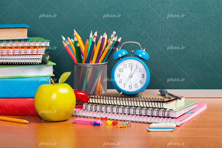 عکس با کیفیت سیب در کنار دفاتر و کتاب های رنگارنگ و ساعت زنگدار بر روی دفاتر و جامداد پر از مداد رنگی بر روی میز چوبی