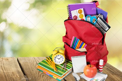 عکس با کیفیت کوله پشتی قرمز و دفتر فنری و آبرنگ و ماشین حساب و قیچی و ماژیک و خط کش در زیپ آن و کتاب های رنگی در کنار آن و ساعت زنگدار بر روی کتاب ها و دفترچه های سفید در کنار آنها و سیب بر روی دفترچه و آنها بر روی میز چوبی