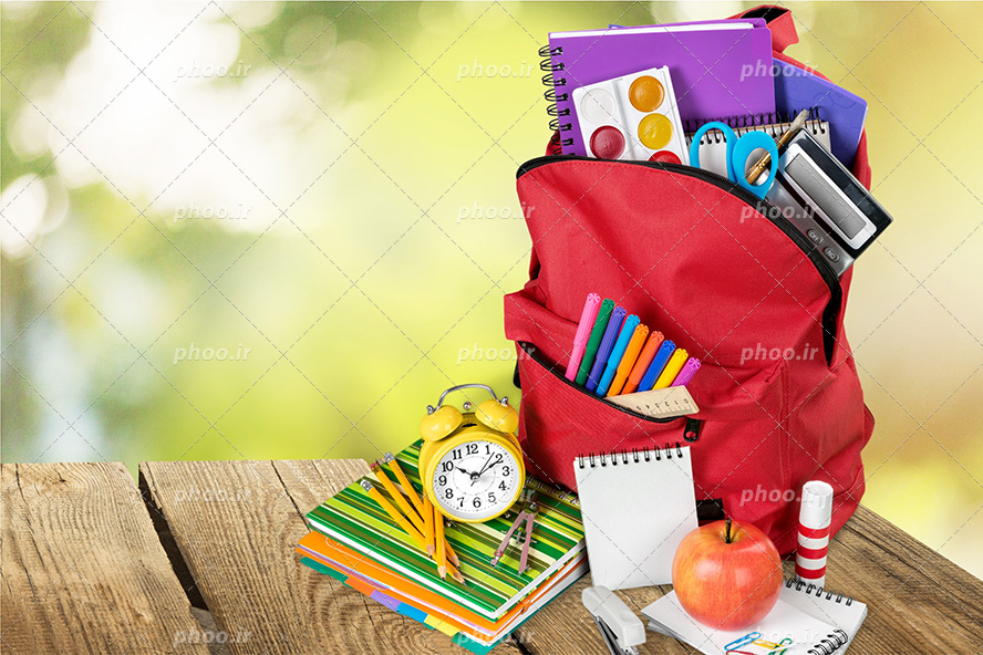 عکس با کیفیت کوله پشتی قرمز و دفتر فنری و آبرنگ و ماشین حساب و قیچی و ماژیک و خط کش در زیپ آن و کتاب های رنگی در کنار آن و ساعت زنگدار بر روی کتاب ها و دفترچه های سفید در کنار آنها و سیب بر روی دفترچه و آنها بر روی میز چوبی