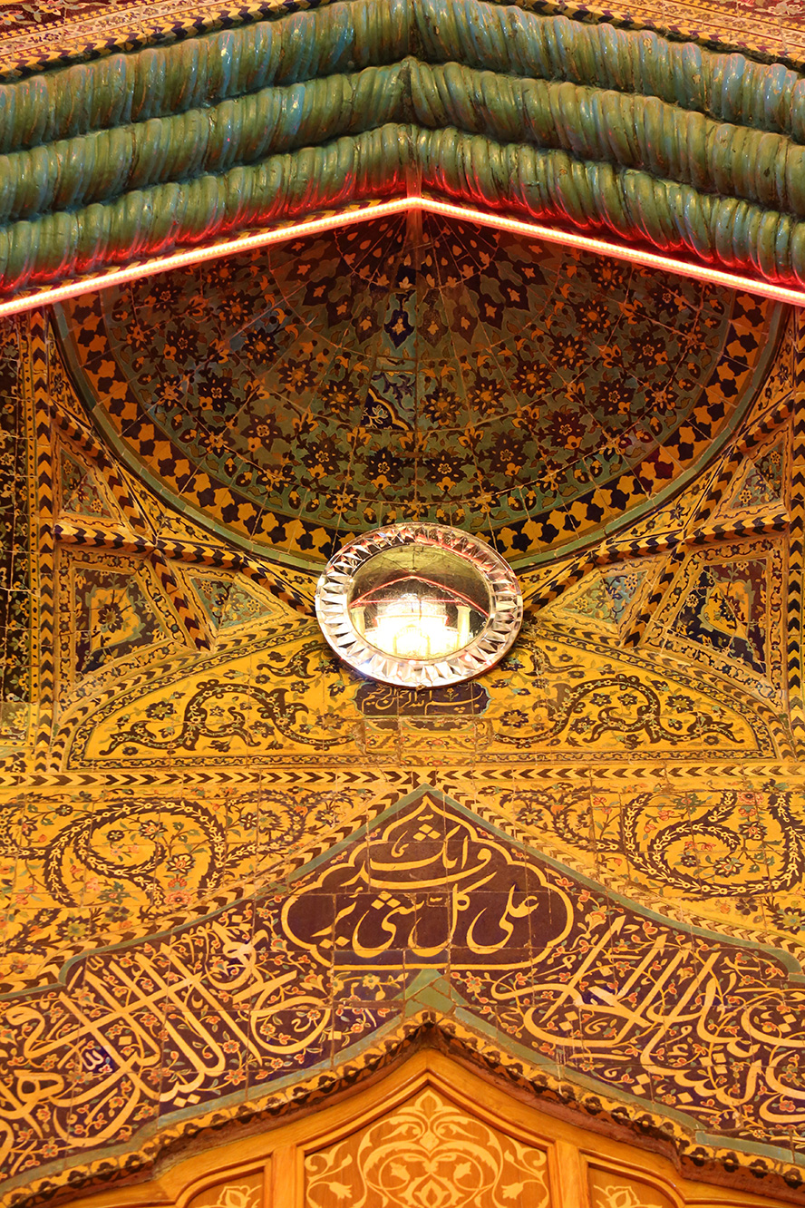 عکس با کیفیت طرح یا پوستر حرم مطهر امام علی (ع) متن انک علی کل شی قدیر بر روی کاشی های دیوار باب الساعه در زیر آینه و نور زرد تابیده بر روی آن