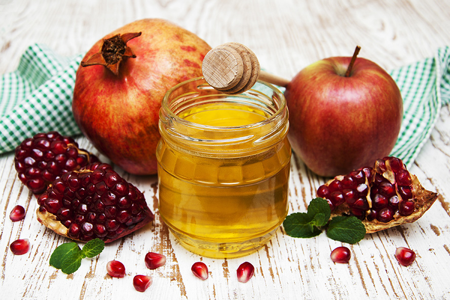 عکس با کیفیت طرح یا پوستر شب یلدا انار در کنار ظرف شیشه ای عسل و قاشق چوبی عسل خوری بر روی آن و سیب در کنار ظرف بر روی میز چوبی و پارچه ای سبز رنگ در پشت آنها
