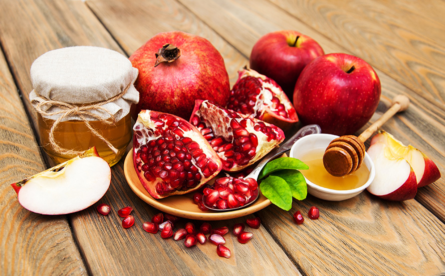 عکس با کیفیت طرح یا پوستر شب یلدا دانه های قرمز انار بر روی قاشق در بشقاب نارنجی و برگ های سبز زیر قاشق در آن اسلایس های انار در کنار ظرف عسل و قاشق عسل خوری بر روی ظرف سفید حاوی عسل و سیب و اسلایس های سیب در کنارش