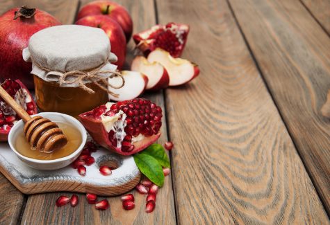عکس با کیفیت طرح یا پوستر شب یلدا تصویر با نور کم و تخته چوبی سفید رنگ و بر روی آن و برگ سبز بر روی آن و در کنار ظرف های عسل و قاشق عسل خوری بر روی ظرف و اسلایس های انار و سیب بر روی تخته