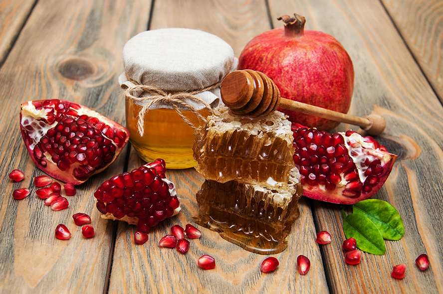 عکس با کیفیت طرح یا پوستر شب یلدا برگ سبز در کنار اسلایس های انار و دو موم عسل بر روی هم و قاشق عسل خوری بر روی موم ها و انار و ظرف عسل در کنار یکدیگر بر روی میز چوبی