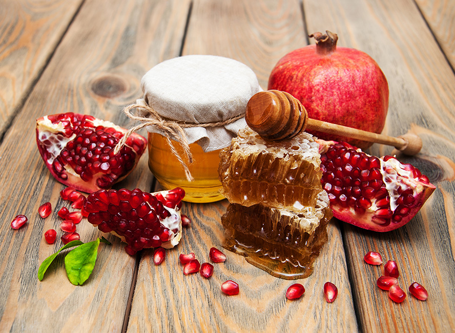 عکس با کیفیت طرح یا پوستر شب یلدا انار و ظرف شیشه ای عسل در کنار یکدگیر و اسلایس ها و دانه های انار در کنار موم های عسل و قاشق چوبی عسل خوری روی موم ها