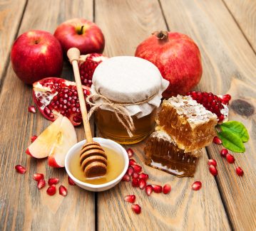 عکس با کیفیت طرح یا پوستر شب یلدا دانه های یاقوتی انار به رنگ قرمز و اسلایس های سیب و انار بر روی میز چوبی در کنار موم های عسل و ظرف سفید و شیشه ای حاوی عسل در وسط و در پشت انار و سیب