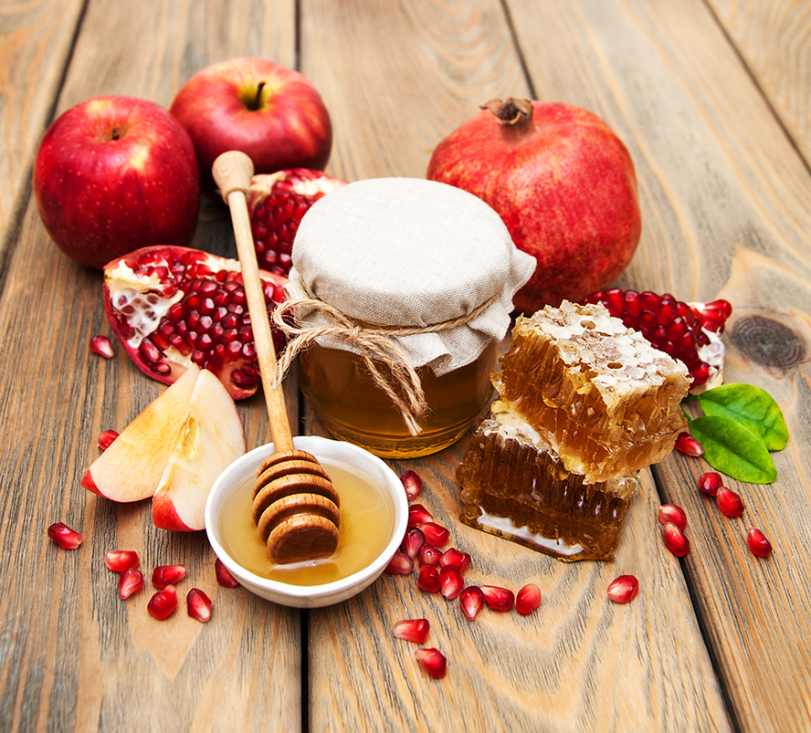 عکس با کیفیت طرح یا پوستر شب یلدا دانه های یاقوتی انار به رنگ قرمز و اسلایس های سیب و انار بر روی میز چوبی در کنار موم های عسل و ظرف سفید و شیشه ای حاوی عسل در وسط و در پشت انار و سیب