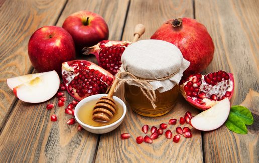 عکس با کیفیت طرح یا پوستر شب یلدا قاشق عسل خوری در ظرف سفید حاوی عسل و اسلایس های سیب و انار در کنارش و دانه های انار ریخته شده بر روی میز چوبی