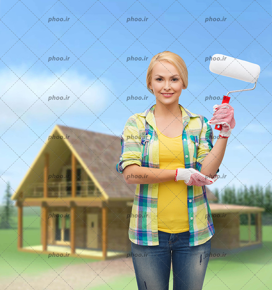 عکس با کیفیت غلتک در دست زن نقاش و در پس زمینه خانه چوبی و آسمان آبی