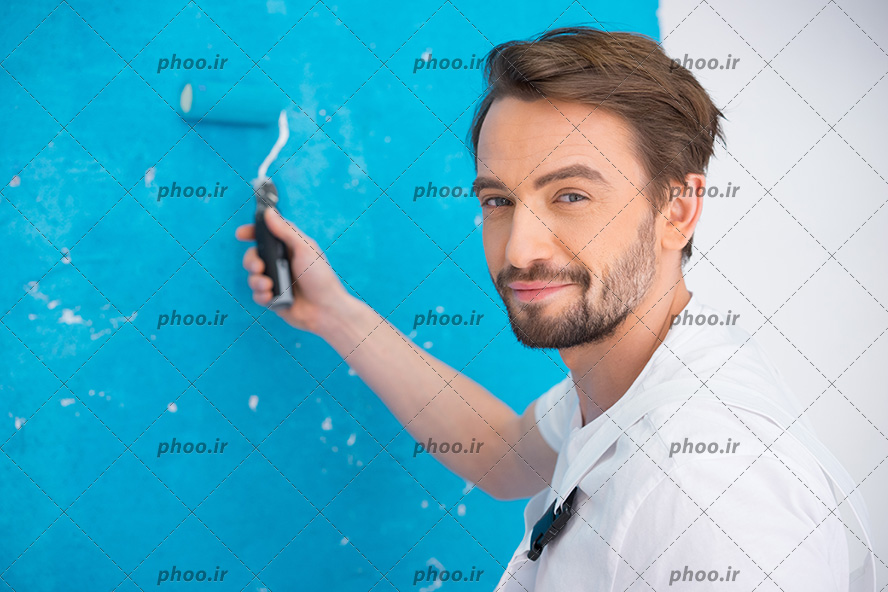 عکس با کیفیت غلتک کوچک در دست مرد جذاب و در حال رنگ آبی زدن به دیوار