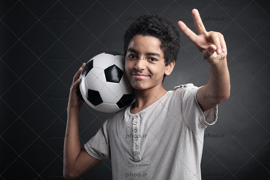 عکس با کیفیت پسر سیاه پوست لبخند به لب و در حال نشان دادن عدد دو با دستش و توپ بر روی شانه او
