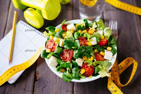 عکس با کیفیت متر اندازه گیری در کنار ظرف سالاد سبزیجات در کنار دفترچه و مداد و دمبل ها