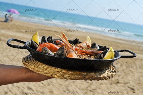عکس با کیفیت غذای دریایی در داخل ماهیتابه و پس زمینه ساحل زیبا