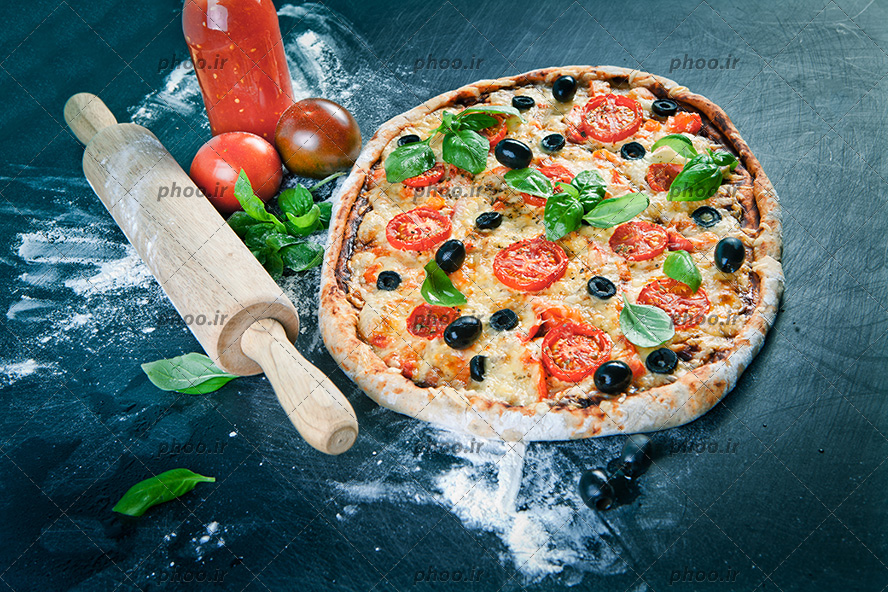 عکس با کیفیت پیتزای خوشمزه تزئین شده با گوجه و سبزیجات و زیتون سیاه در کنار وردنه و سس کچاپ و دو عدد گوجه