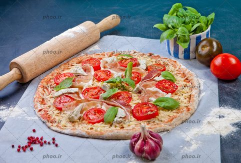 عکس با کیفیت وردنه در کنار پیتزا خوشمزه تزئین شده با گوجه و پپرونی و سبزیجات و سیر و گوجه و مقداری ریحان در کنار پیتزا