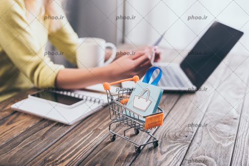 عکس با کیفیت خرید ها داخل سبد چرخدار کوچک بر روی میز در کنار مرد در حال کار کردن با لپ تاپ