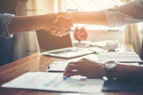 عکس با کیفیت دو مرد در حال دست دادن به یکدیگر و لپ تاپ و کاغذ بر روی میز کار