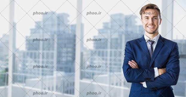 عکس با کیفیت مهندس با کت و شلوار سرمه ای و لبخند بر لب ایستاده در مقابل پنجره و ساختمان های در حال ساخت