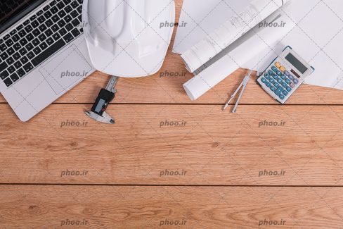 عکس با کیفیت لپ تاپ در کنار کلاه ایمنی به رنگ سفید در کنار نقشه های ساختمان و ماشین حساب