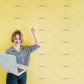 عکس با کیفیت لپ تاپ در دست زن با چهره ی خندان و در حال اشاره به بالای سر و پس زمینه دیوار زرد