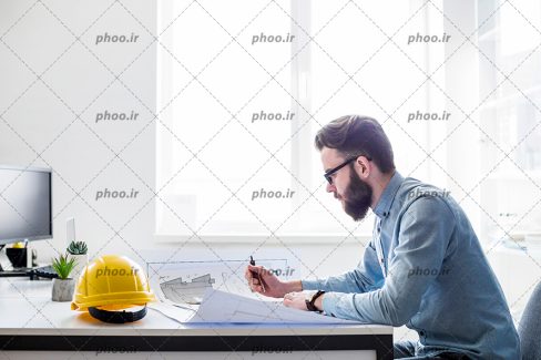 عکس با کیفیت مرد درحال دیدن نقشه روی میز و کلاه ایمنی در کنارش و مانیتور در رو به روی متن