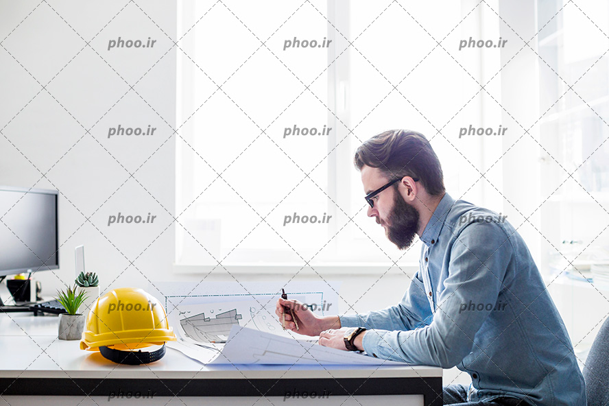 عکس با کیفیت مرد درحال دیدن نقشه روی میز و کلاه ایمنی در کنارش و مانیتور در رو به روی متن