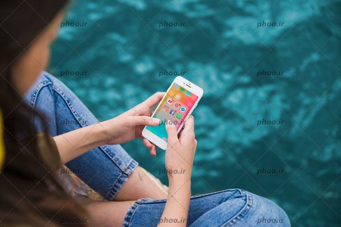 عکس با کیفیت دختر نشسته در کنار استخر و گوشی موبایل در دستانش و اینستاگرام و فیش بوک و واتس آپ و یوتیوب در صغحه نمایش گوشی