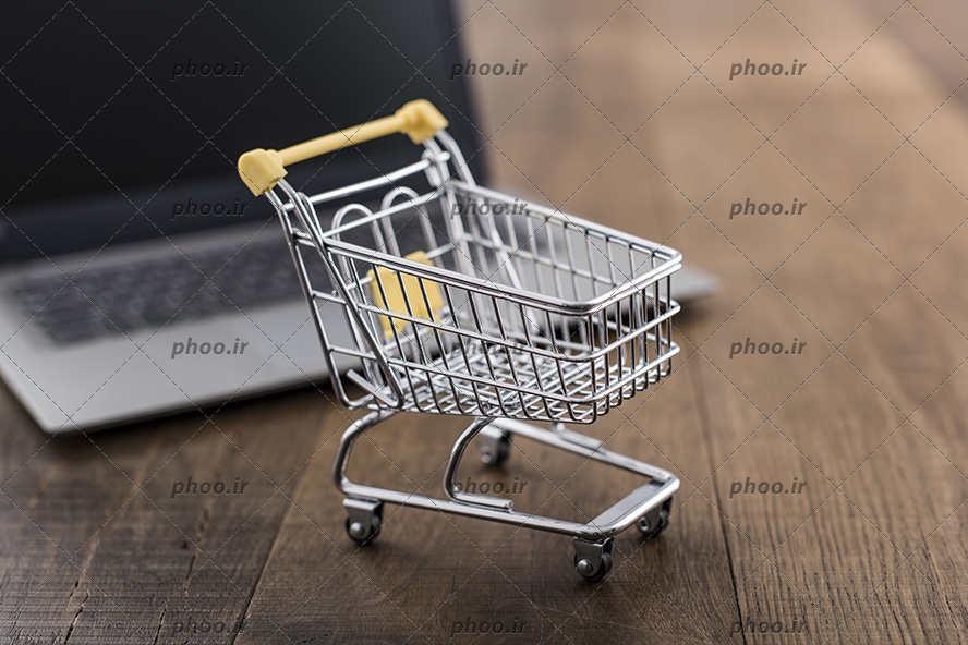 عکس با کیفیت سبد خرید چرخدار کوچک با دسته های زرد بر روی میز چوبی و در پشت آن لپ تاپ