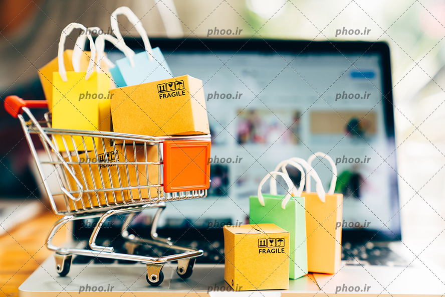 عکس با کیفیت ماکت زیبای سبد خرید چرخدار و بسته بندی ها و پاکت های زیبا در آن بر روی لپ تاپ
